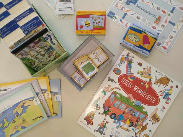 Verschiedene Materialien für die Sprachförderung liegen nebeneinander: 4 Lesehefte, eine Box mit Bildkarten zu verscheidenen Alltagsbereichen wie Schule, Küche und Spielen getrennt durch Karteireiter, Memory-Karten, ein Wimmelbilderbuch, zwei weitere klei