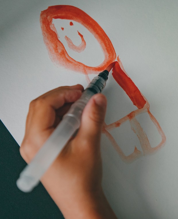 Eine Kinderhand hält einen Pinselstift. das Kind malt einen roten menschen auf das Papier.