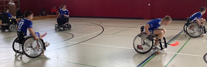 mehrere Personen in blauen Trikots fahren in Sportrollstühlen mit Hockeyschlägern in einer Halle