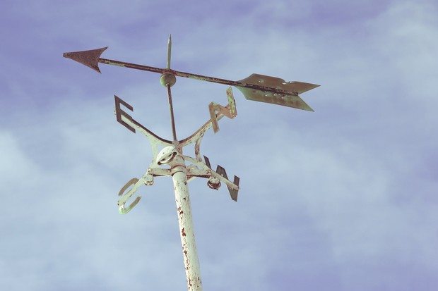 Ein weißer Windrichtungsanzeiger aus Metall vor blauem Himmel: ein Kreuz, aden vier Enden die Anfangsbuchstaben der Himmelsrichtung, darüber ein Pfeil, der  Richtung Süd-Ost weist.