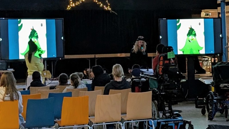 Zwei Frauen sprechen in ein Mikrophon,im Hintergrund leuchten Bildschirme mit Weihnachtsbäumen. Viele Zuschauer schauen zu.
