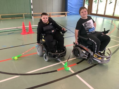 Zwei Jungen im Elektro-Rollstuhl mit je einem Hockeyschläger in je einer Hand stehen in einer Turnhalle und lächeln in die Kamera.